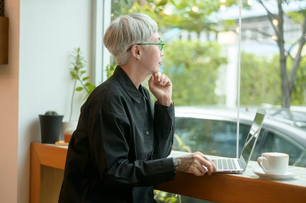 Executive woman sitting at a laptop and looking out the window, reflecting on courageous leadership decisions and the importance of executive courage.