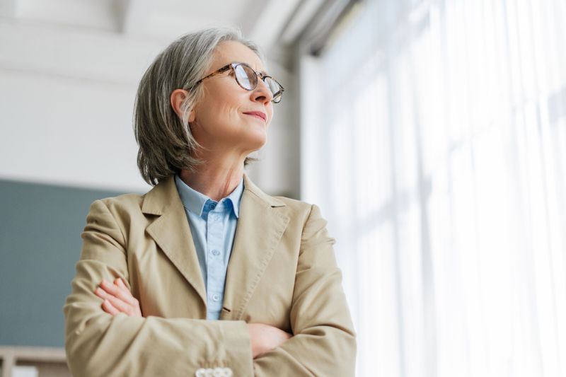 Professional woman standing near a window, reflecting calmly with soft natural light symbolising balance and renewal.