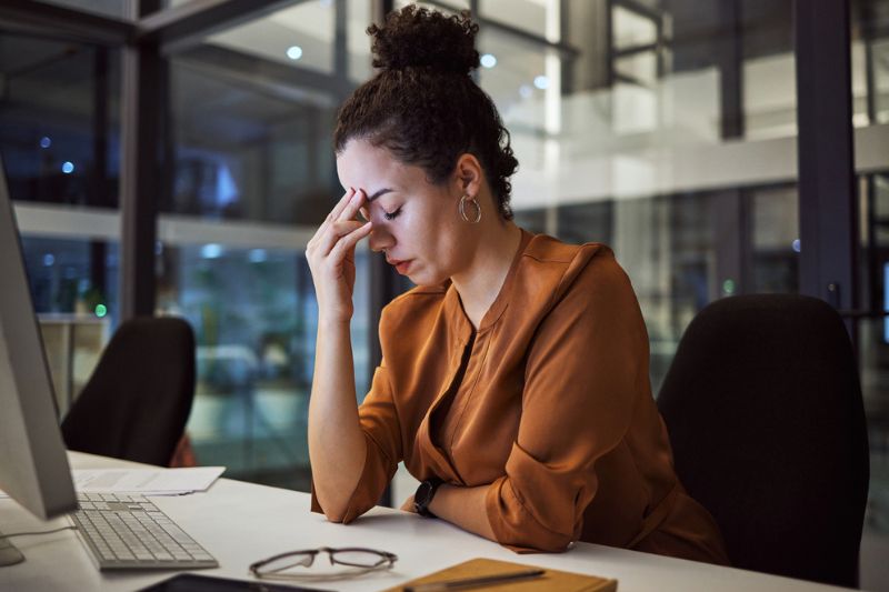 Professional woman sitting at her desk late in the office, pausing with her hand on her forehead — symbolising the warning signs of burnout in leaders and teams.